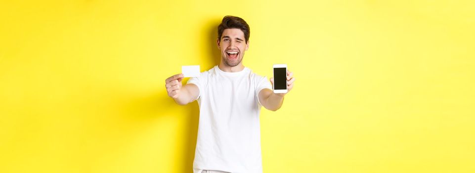 Happy man showing good online offer on mobile phone screen, holding credit card and winking, standing over yellow background.