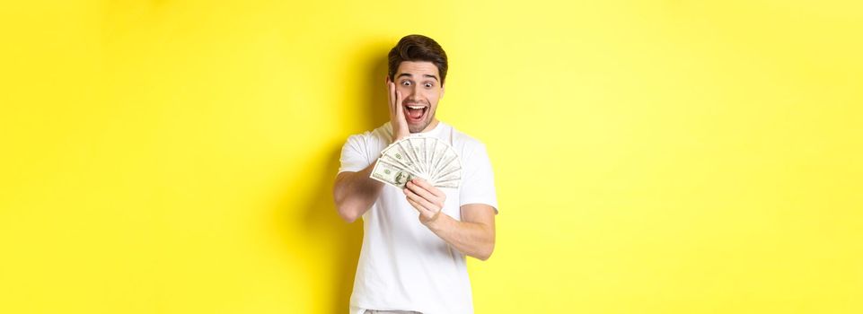 Man looking amazed at money, winning cash prize, standing against yellow background.