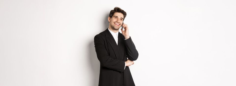 Image of handsome and successful businessman talking on phone, smiling pleased, standing in suit against white background.