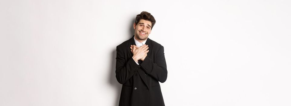 Portrait of happy and pleased handsome man in party suit, holding hands on heart and sighing flattered, saying thank you, standing grateful over white background.