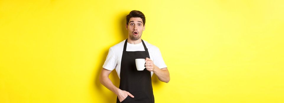 Barista holding coffee mug and looking surprised, standing in black apron cafe uniform against yellow background.