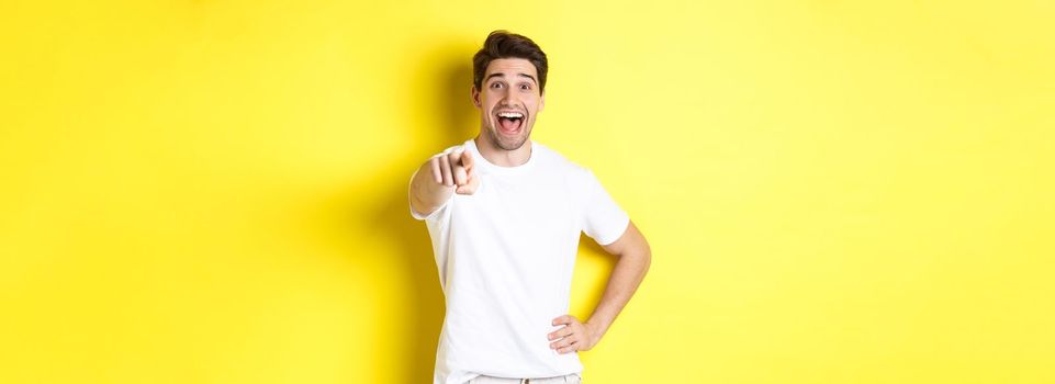 Happy guy pointing finger at camera and laughing, check out something, standing over yellow background.