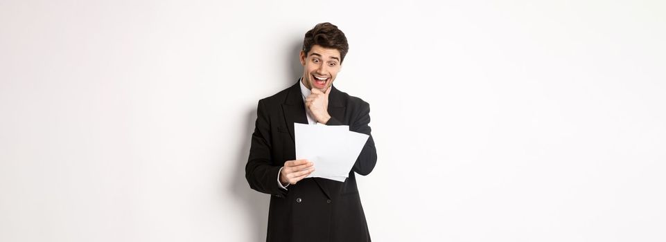 Portrait of handsome businessman looking excited at documents, working, standing against white background in black suit.