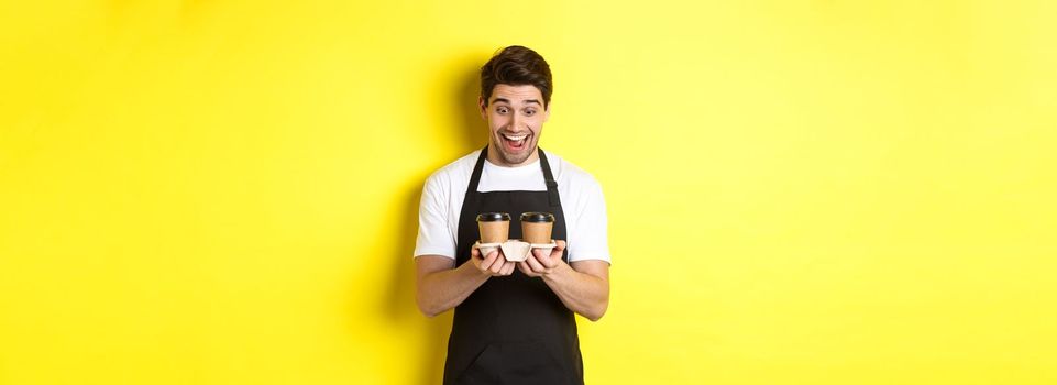 Waiter looking excited at two cups of takeaway coffee, wearing black apron, standing over yellow background.