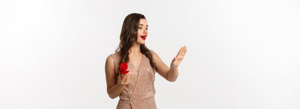 Beautiful woman in luxury dress, trying on engagement ring, saying yes to marriage proposal, looking dreamy at hand, standing over white background.