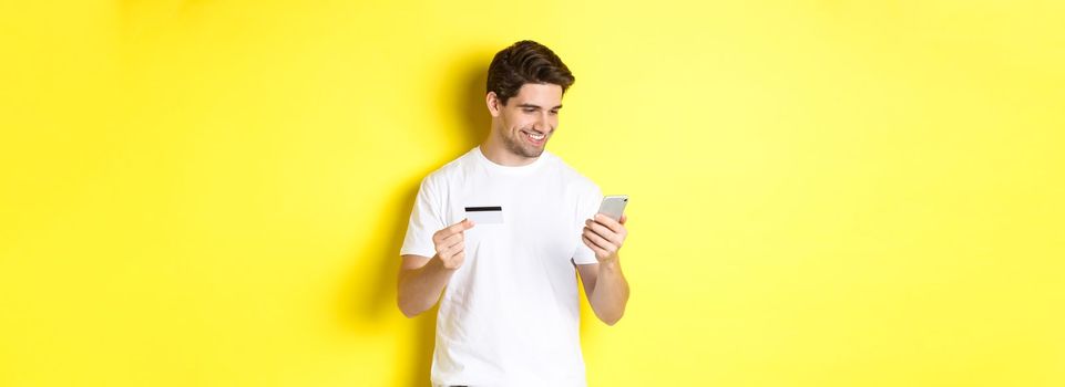 Young man paying online, insert credit card number on mobile phone, shopping in internet, standing over yellow background.