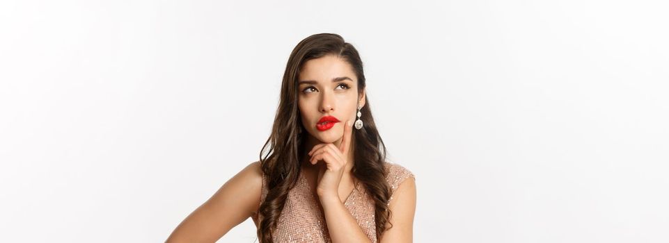 Concept of New Year celebration and winter holidays. Close-up of elegant woman with red lips and dress, looking at upper left corner and thinking, standing over white background.