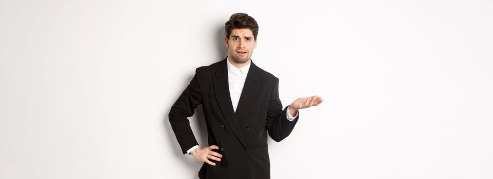 Image of confused handsome man in formal suit, raising hand and shrugging, cant understand something, standing against white background.