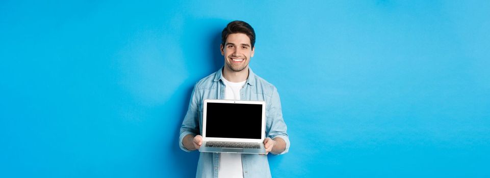 Handsome young man introduce product on laptop screen, showing computer and smiling, standing over blue background.