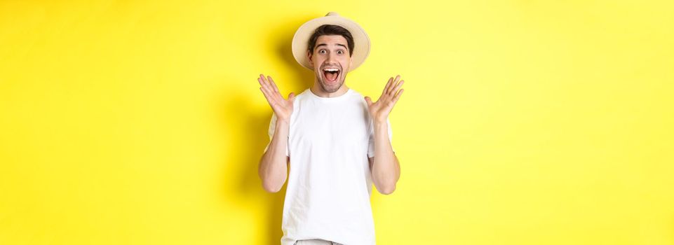 Concept of tourism and summer. Happy young man in straw hat looking amazed, reacting to surprise, standing over yellow background.