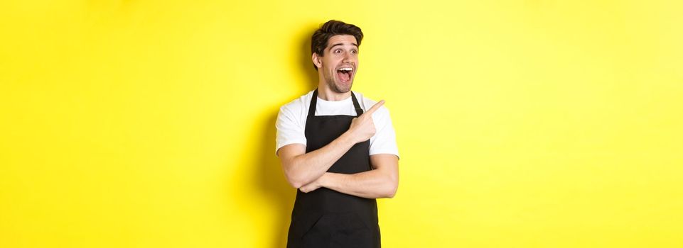 Waiter in black apron checking out promo offer, pointing finger and looking left at your logo, standing over yellow background.