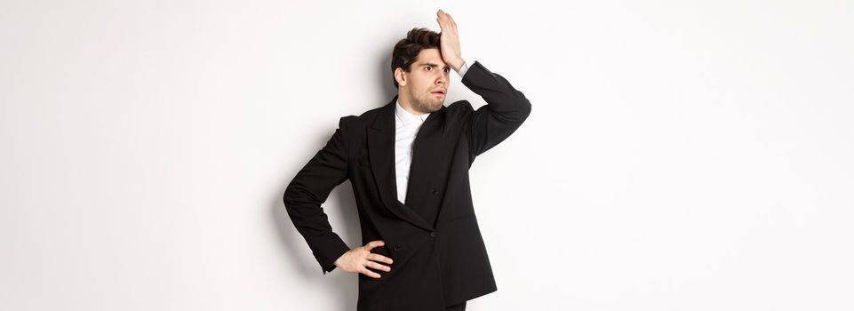 Portrait of shocked and distressed businessman in black suit, slap forehead and looking anxious, standing troubled over white background.