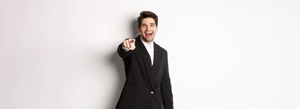 Concept of new year party, celebration and lifestyle. Portrait of handsome stylish man in black suit, smiling and pointing finger at camera, standing over white background.