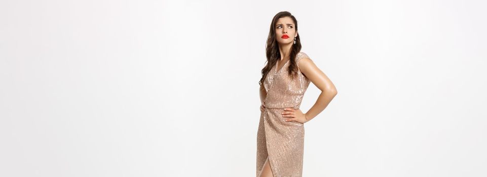 Christmas party and celebration concept. Full length of jealous woman looking left with displeased face, frowning and staring aside, standing in glamour dress, white background.