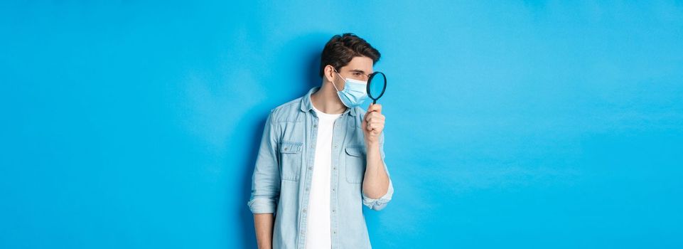 Concept of coronavirus, social distancing and pandemic. Man in medical mask searching for something, looking left through magnifying glass, studying copy space, blue background.