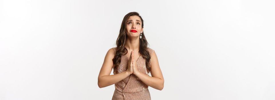 Celebration and party concept. Hopeful young woman begging for help, looking up and praying, pleading God, standing in glamour dress over white background.