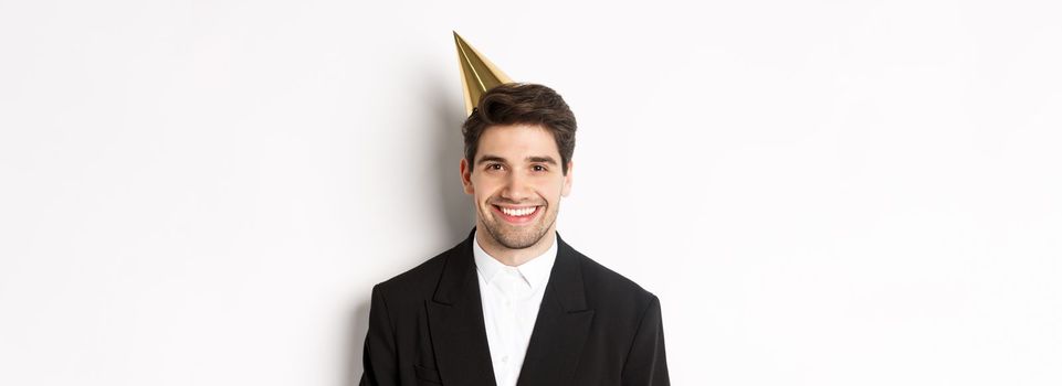 Close-up of handsome happy man in suit and party hat, smiling joyful, celebrating holiday, standing against white background.