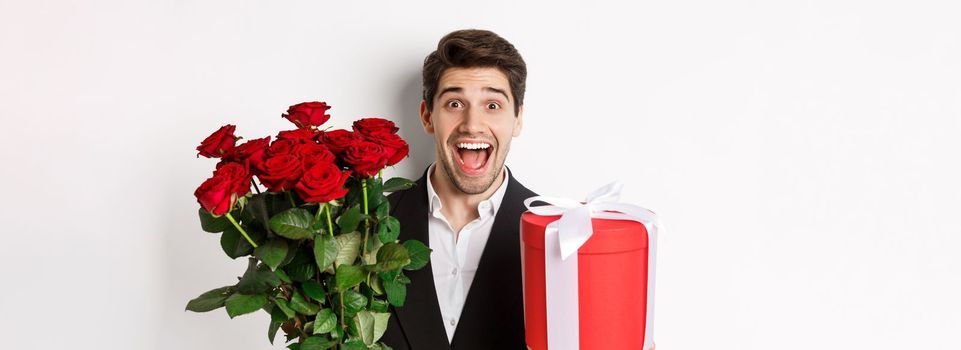 Close-up of handsome bearded man in suit, holding present and bouquet of red roses, smiling at camera, standing against white background.