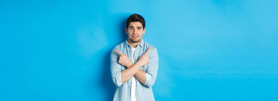 Portrait of nervous guy pointing fingers sideways, looking indecisive and asking for help with choice, showing left and right promo offers, standing against blue background.