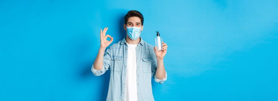 Concept of covid-19, pandemic and social distancing. Satisfied young man in medical mask recommending hand sanitizer, showing ok sign and antiseptic, standing against blue background.