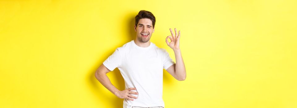 Confident handsome man winking, showing okay sign in approval, like something good, standing over yellow background.