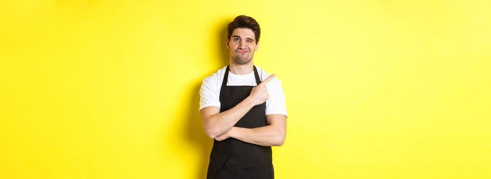 Displeased barista in black apron pointing finger at upper left corner, grimacing skeptical, dislike something, standing against yellow background.