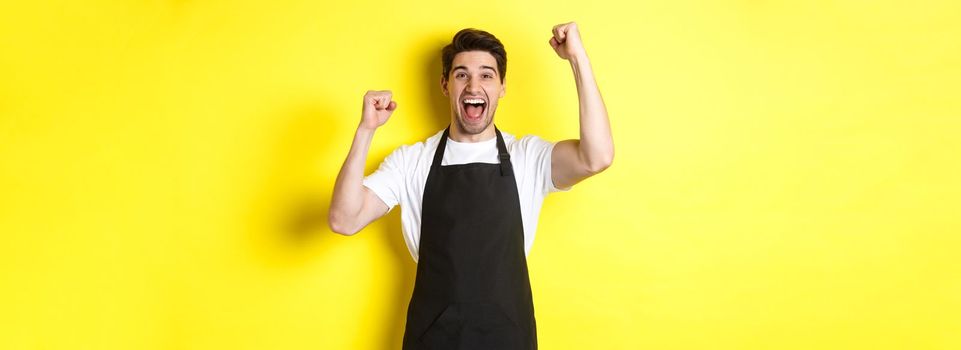 Happy barista celebrating victory, raising hands up and shouting for joy, wearing black apron, shop uniform, standing against yellow background.