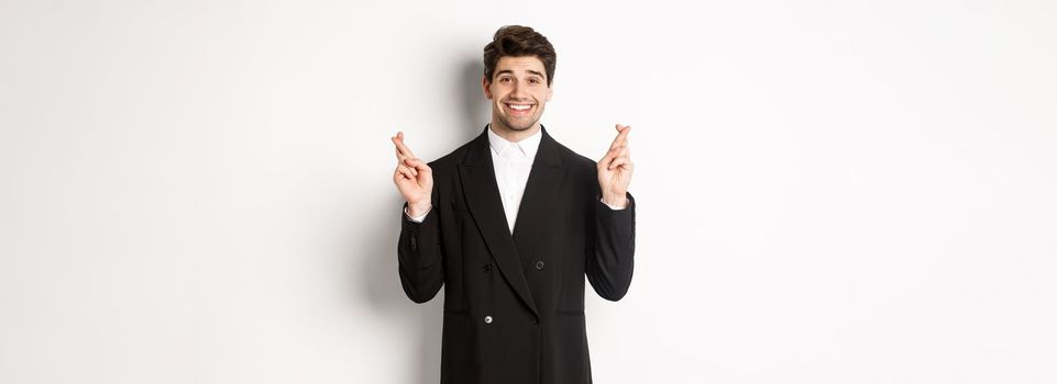 Hopeful and optimistic young businessman in suit, smiling while crossing fingers for good luck and making wish, standing over white background.