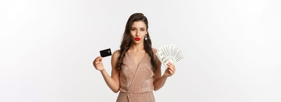 Shopping concept. Sassy woman in elegant dress showing credit card with money, looking pleased at camera, standing over white background. Copy space