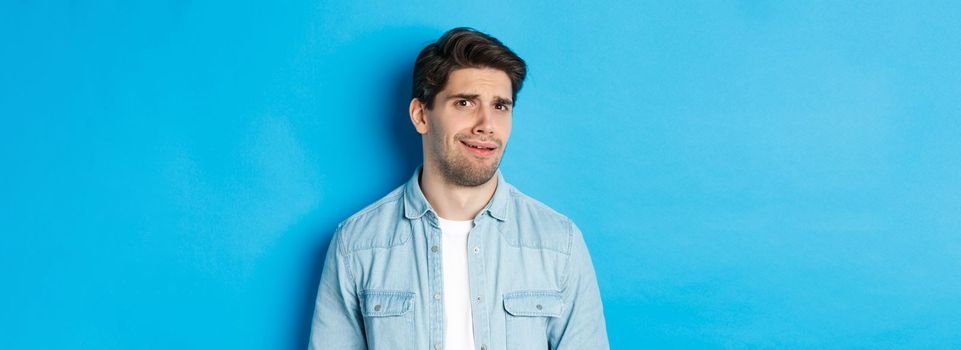 Confused and uncomfortable man looking at something strange or creepy, cringe from bad advertisement, standing over blue background.