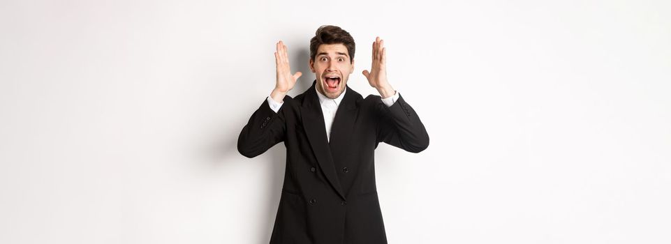 Frustrated and concerned man in black suit, screaming in panic and looking at something shocking, standing over white background.