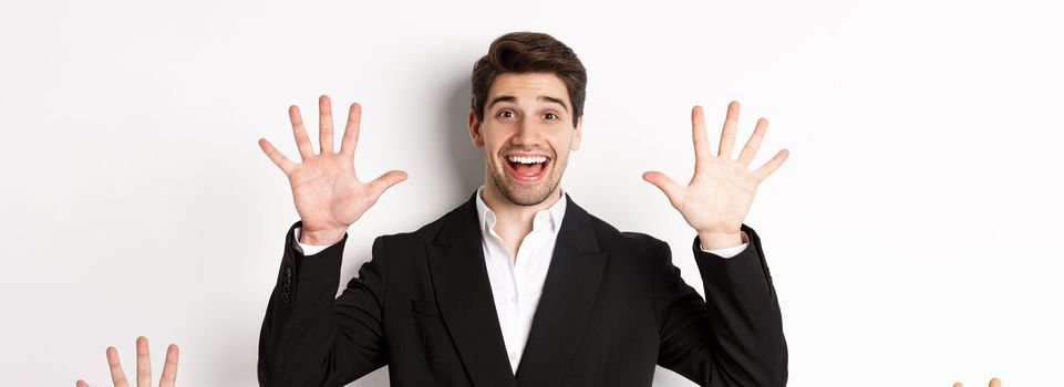 Close-up of handsome businessman in black suit, smiling amazed, showing number ten, standing over white background.