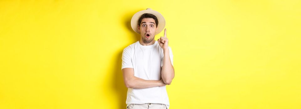 Portrait of young man in straw hat having an idea, raising finger eureka sign, making suggestion, standing over yellow background.