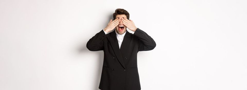 Image of handsome stylish man in black suit, waiting for christmas surprise, covering eyes with hands and smiling, anticipating presents, standing over white background.
