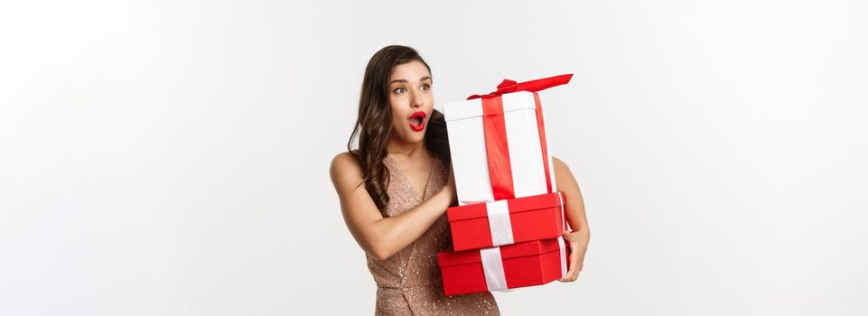 New Year, Christmas and celebration concept. Image of beautiful woman in luxury dress, holding holiday gifts and looking left surprised, standing over white background.