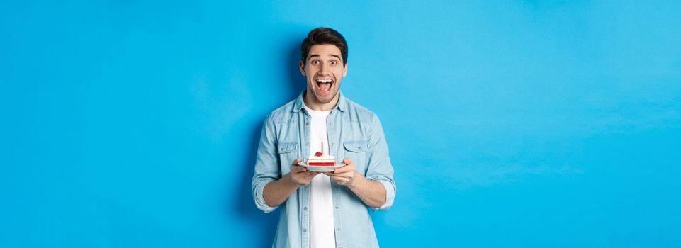 Excited guy holding birthday cake and smiling at camera, celebrating b-day over blue background.