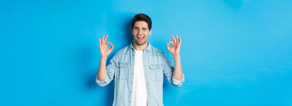 Relaxed and confident man showing ok signs and winking, everything okay gesture, standing against blue background.