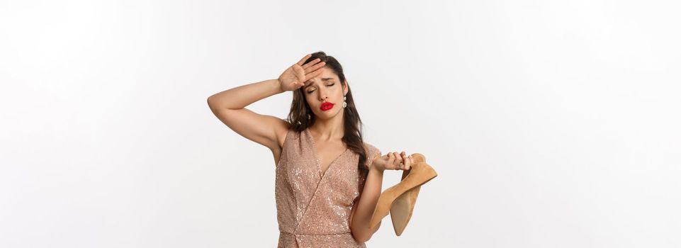 Party and celebration concept. Tired woman in elegant dress take off pair of heels, holding hand on forehead, express fatigue, standing over white background.