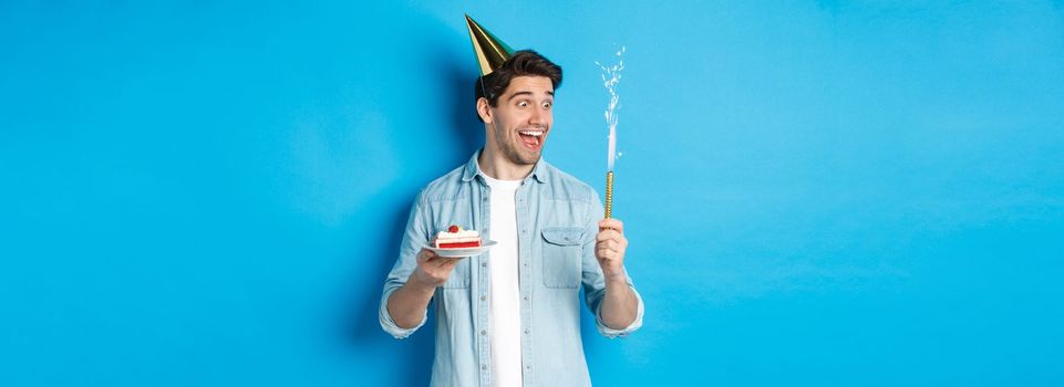 Happy young man celebrating birthday in party hat, holding b-day cake and smiling, standing over blue background.