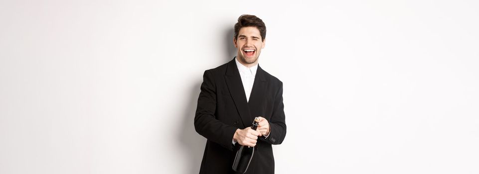 Image of handsome, confident man in black suit, celebrating holiday, open a bottle of champagne and partying, standing joyful against white background.