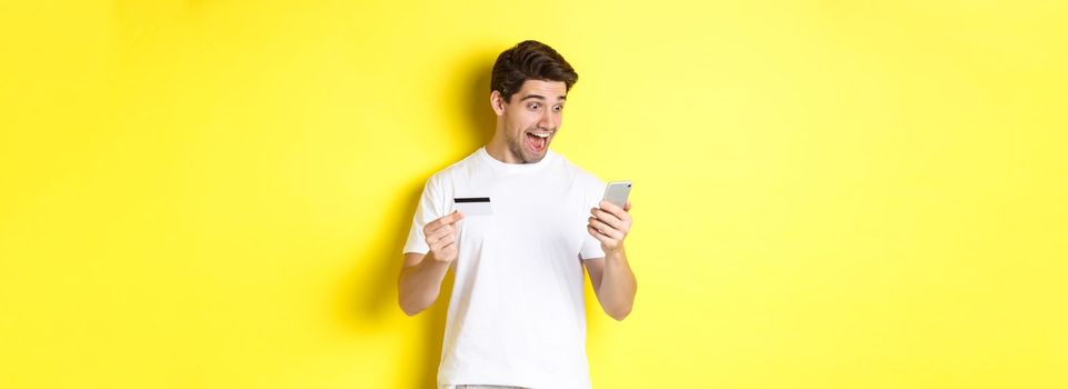 Man looking surprised at smartphone, shopping online, holding credit card, standing over yellow background. Copy space