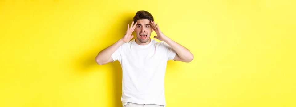 Frustrated guy looking shocked, panicking and holding hands on head, standing against yellow background.