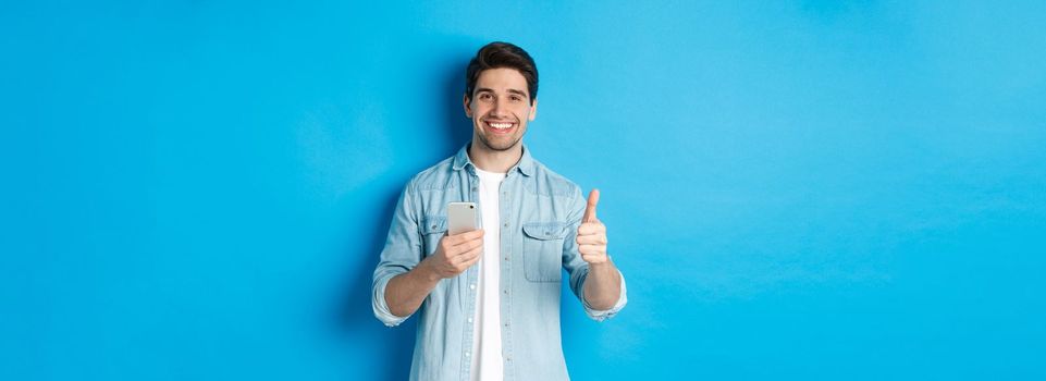 Concept of online shopping, applications and technology. Satisfied man in casual clothes smiling, showing thumbs up after using smartphone app, standing over blue background.