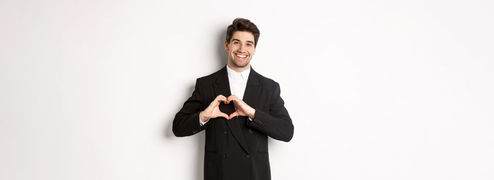 Image of handsome groom in black suit, showing heart sign and smiling, express love and sympathy, standing over white background.