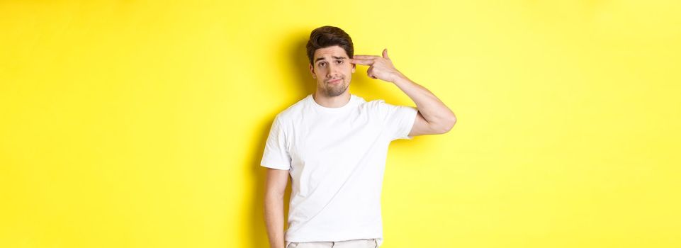 Bothered man shooting himself with finger gun, looking displeased and tired, standing against yellow background.