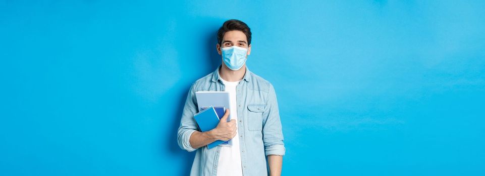 Education, covid-19 and social distancing. Guy student in medical mask looking happy, holding notebooks, standing over blue background.