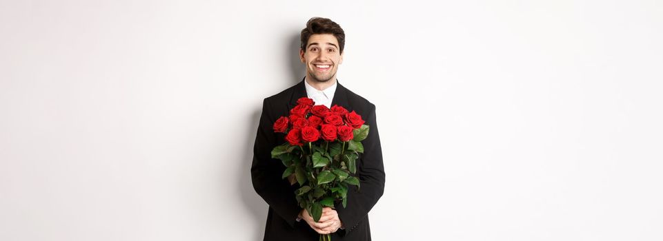 Image of handsome man in black suit, holding bouquet of roses and smiling, standing against white background.