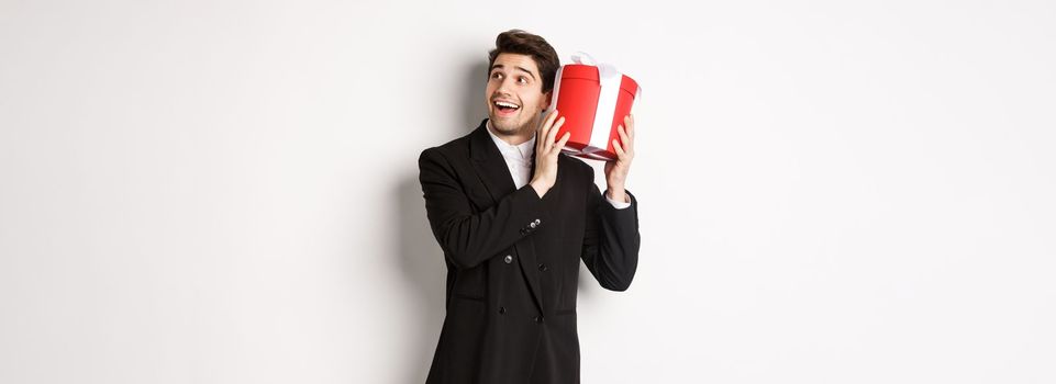 Concept of christmas holidays, celebration and lifestyle. Image of attractive man in black suit, shaking present to guess what inside, enjoying new year, standing against white background.