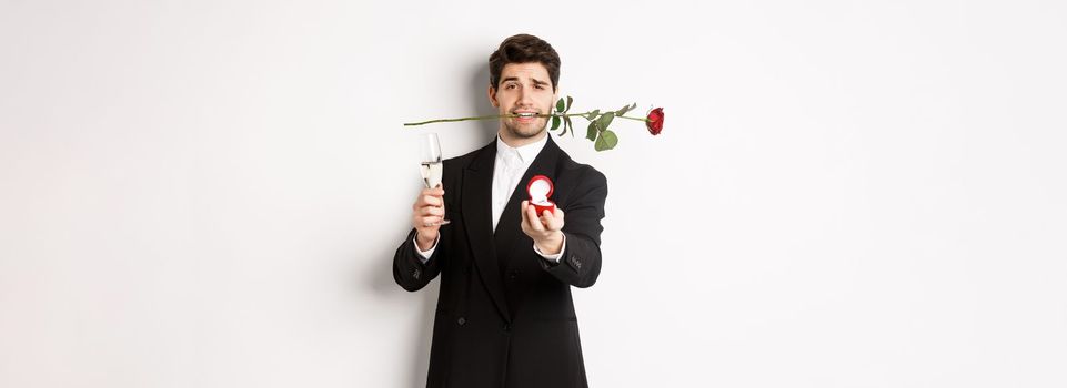 Romantic young man in suit making a proposal, holding rose in teeth and glass of champagne, showing engagement ring, asking to marry him, standing against white background.