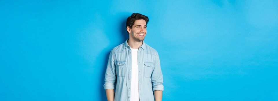 Handsome smiling adult man in casual outfit, smiling and looking left at promo offer, standing against blue background.